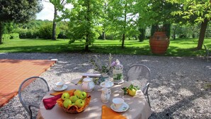 Een buitentafel gedekt met een mand appels, kopjes en een theepot, omringd door stoelen en een grote plantenbak.