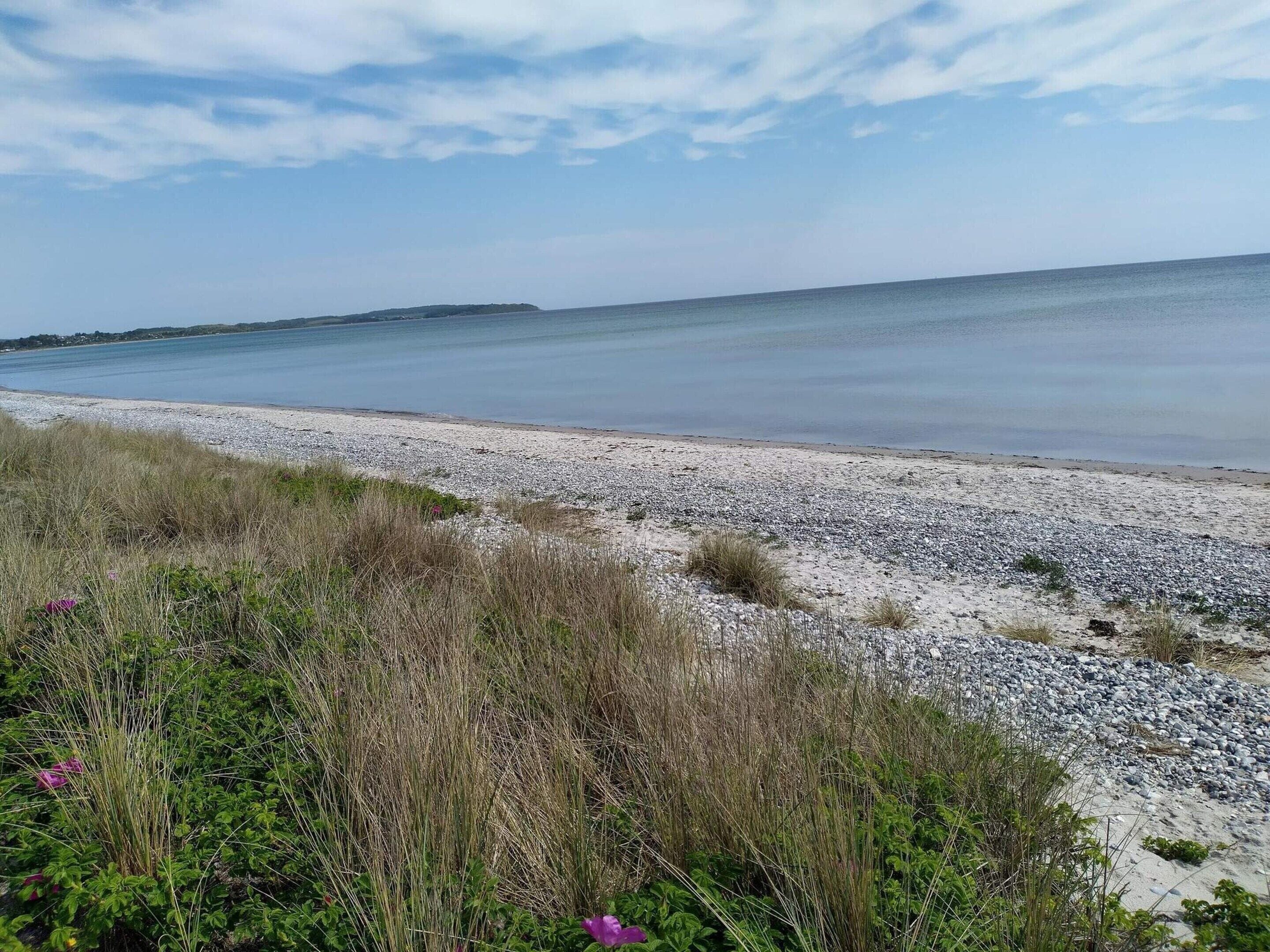 Ein Strand mit Kieselsteinen und Gras im Vordergrund, ein ruhiges Meer und ein klarer Himmel.