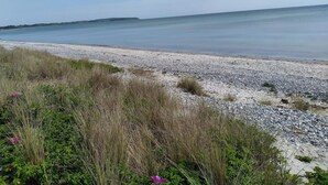 Ein Strand mit Kieselsteinen und Gras im Vordergrund, ein ruhiges Meer und ein klarer Himmel.
