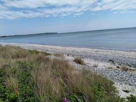 Una playa con piedras y hierba en primer plano, un mar tranquilo y un cielo despejado.