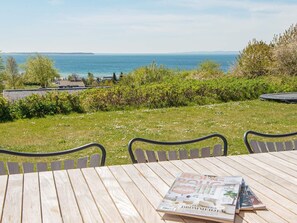 A wooden table with chairs, a magazine, and a view of the sea and distant buildings. - 5 Star Holiday Home in Ebeltoft (Ebeltoft)