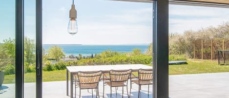 A dining area with a table and chairs, a hanging light fixture, and a view of the sea through large glass doors.