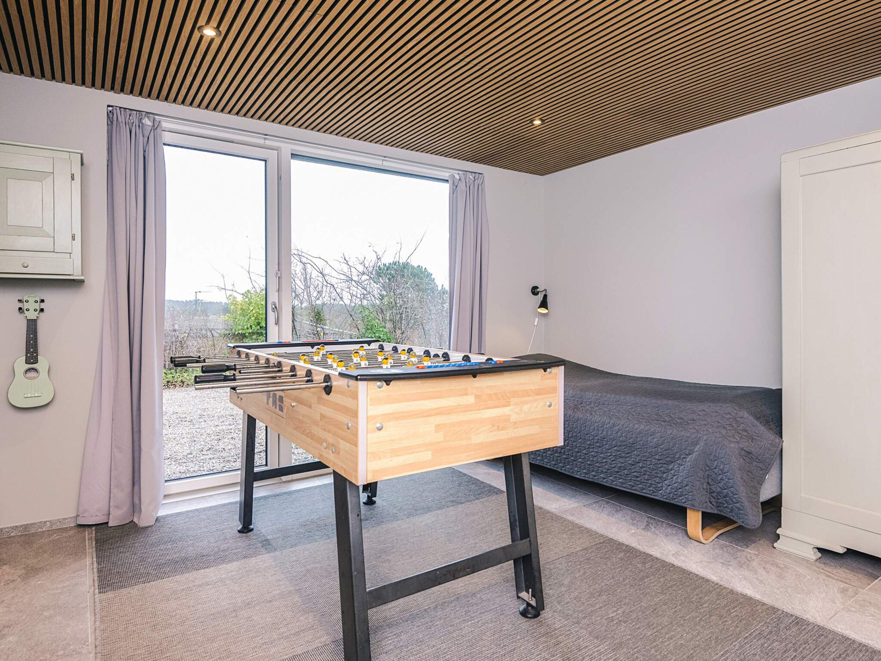 A room with a wooden foosball table, a bed, a guitar, and a window with curtains.