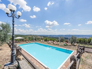 A rectangular swimming pool with a stone border, surrounded by lounge chairs and umbrellas, set against a backdrop of rolling hills and a clear sky. - Belvilla by OYO Istrice (Capraia e Limite)