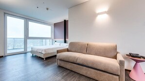 A modern hotel room with a large bed, a sofa, a small pink table, and a large window offering a city view. - Tranquil Apartment in Venice near Bosco Dell'ossilleno Park (Mestre)