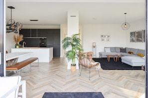 A modern living space with a herringbone floor, a dining area with a bench, a sofa set, a potted plant, and a kitchen in the background.