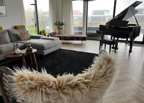 A living room with a grand piano, a grey sofa, a black rug, and a fluffy white chair.