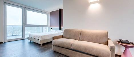 A modern hotel room with a large bed, a sofa, a small pink table, and a large window offering a city view.