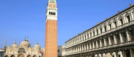 A historic square with a prominent clock tower, a grand building with classical architecture, and a bustling crowd.