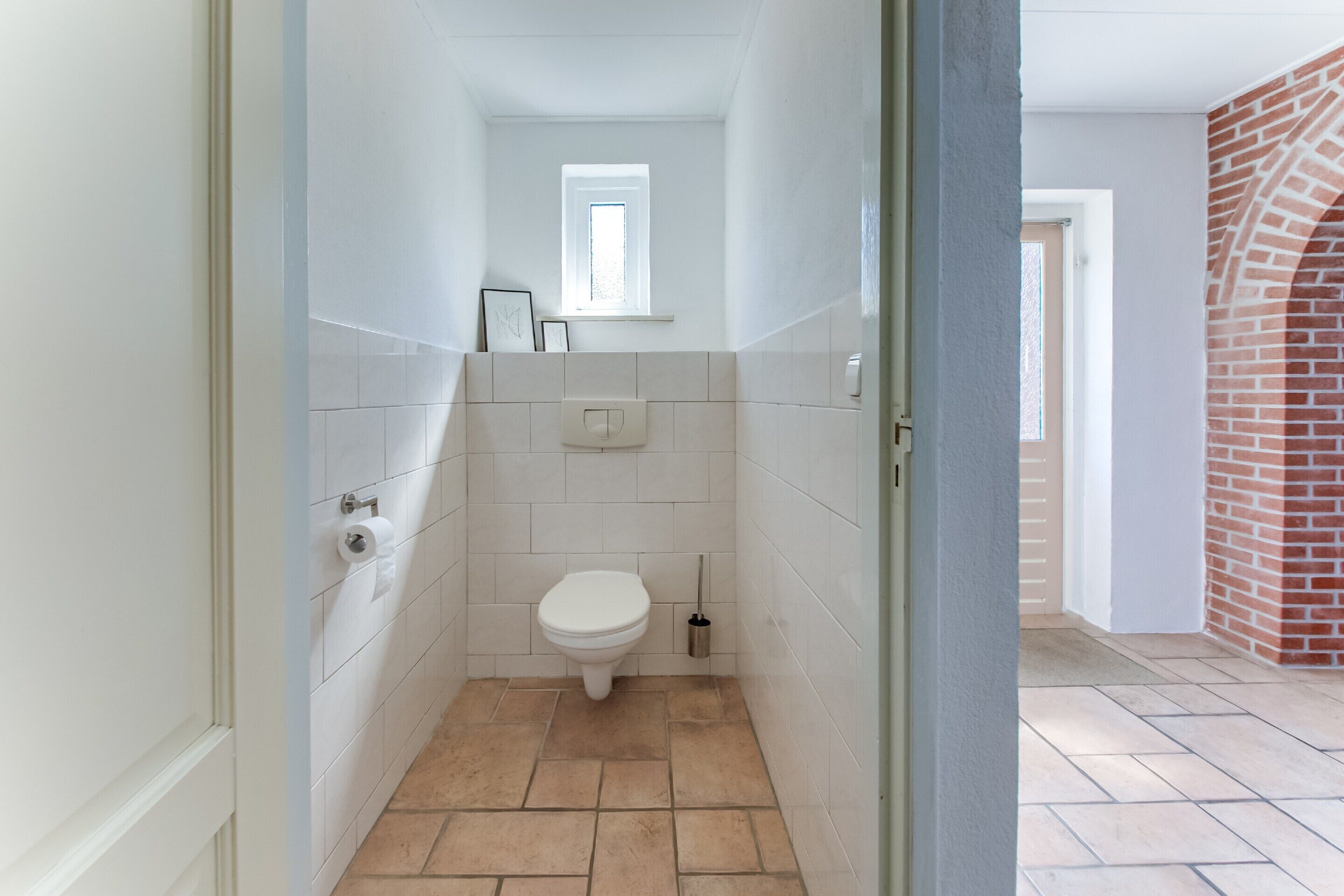 A narrow bathroom with white tiles, a white toilet, a small window, and a door leading to a hallway with a brick wall.
