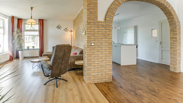 Un vaste espace de vie doté d’un plancher en bois, d’un fauteuil en cuir marron, d’une table basse ronde et d’un canapé. On y trouve également une colonne en briques et une arche qui mène à la cuisine.