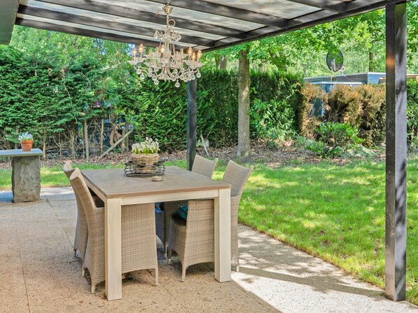 An outdoor patio with a wooden table and wicker chairs, a hanging chandelier, and a stone planter with white flowers.