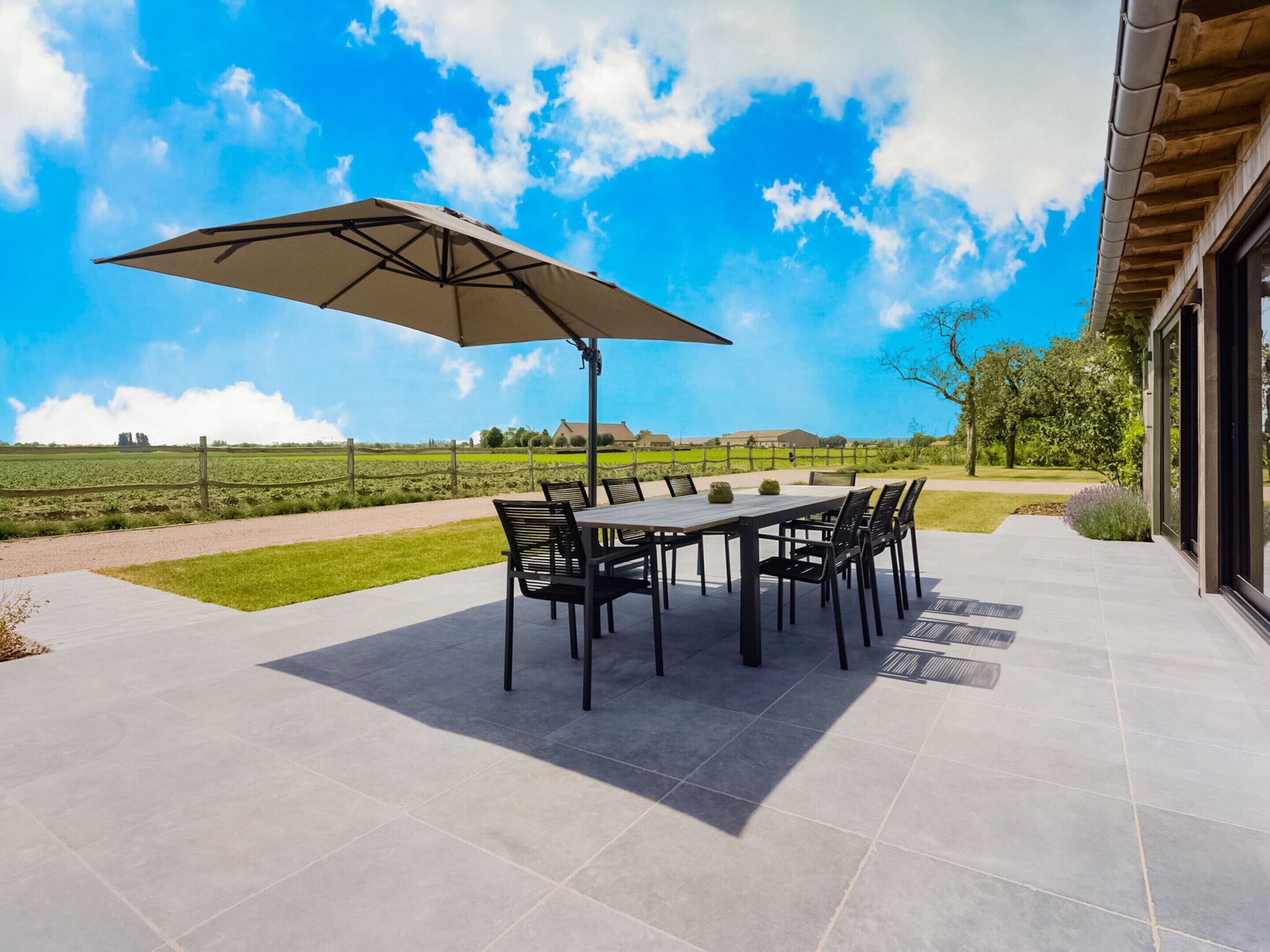 Une terrasse extérieure avec une table et des chaises, un grand parasol et une vue sur un champ verdoyant et des arbres.