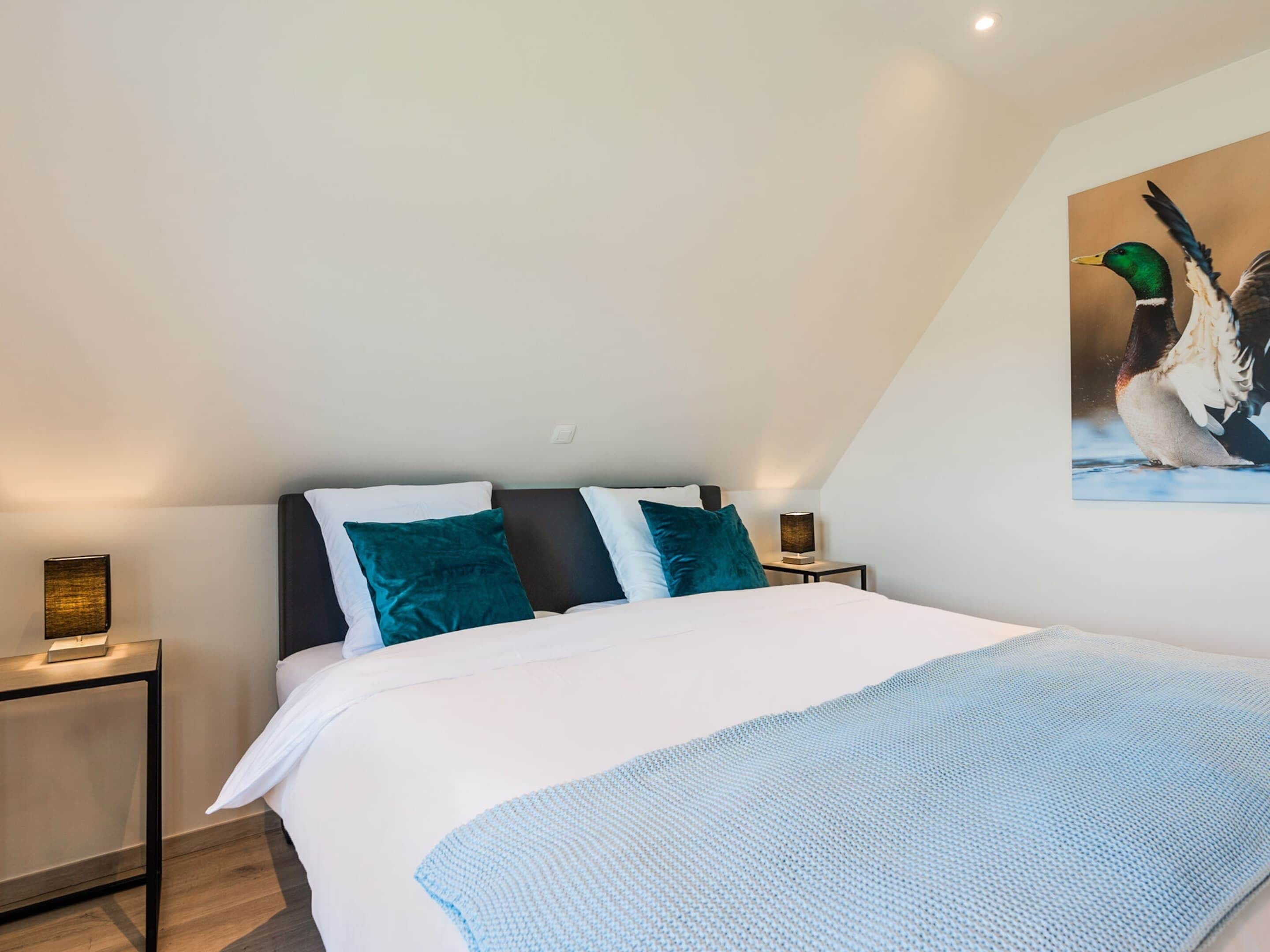 A bedroom with a bed, pillows, a nightstand, a lamp, and a wall-mounted painting of a duck.