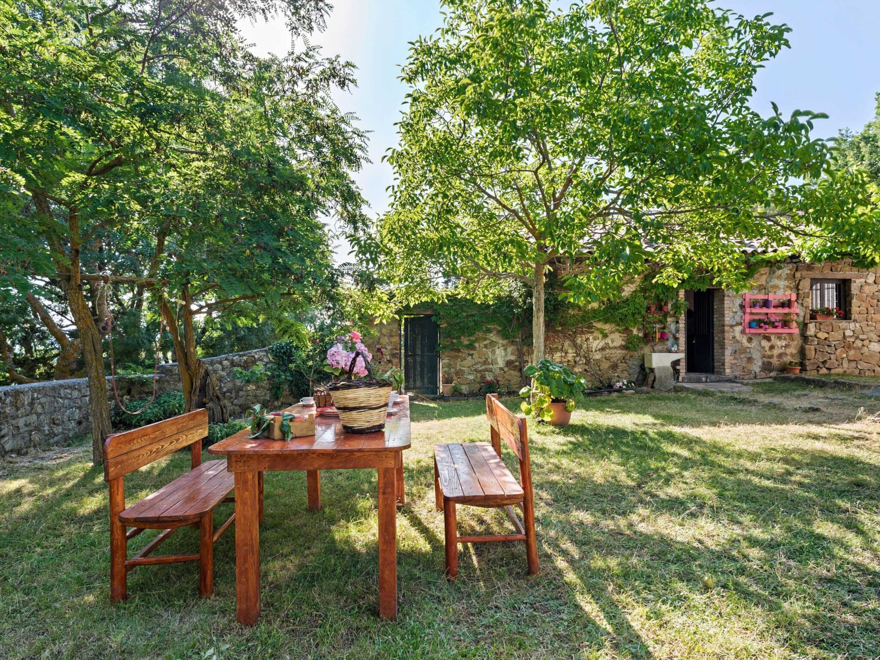 An outdoor seating area with a wooden table and benches, surrounded by lush greenery and a stone building.