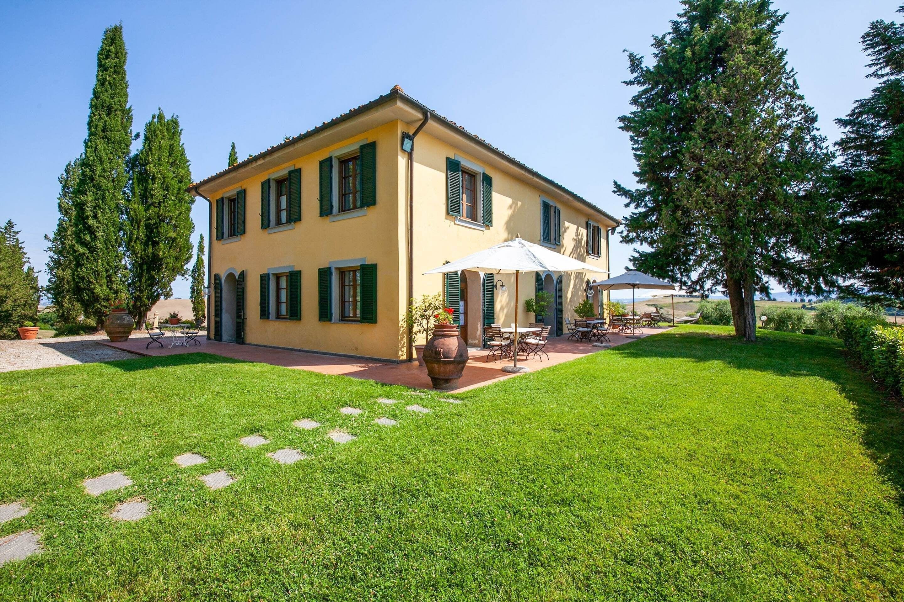 Ein gelbes Gebäude mit grünen Fensterläden, eine Terrasse mit Gartenmöbeln und einem gepflegten Rasen.