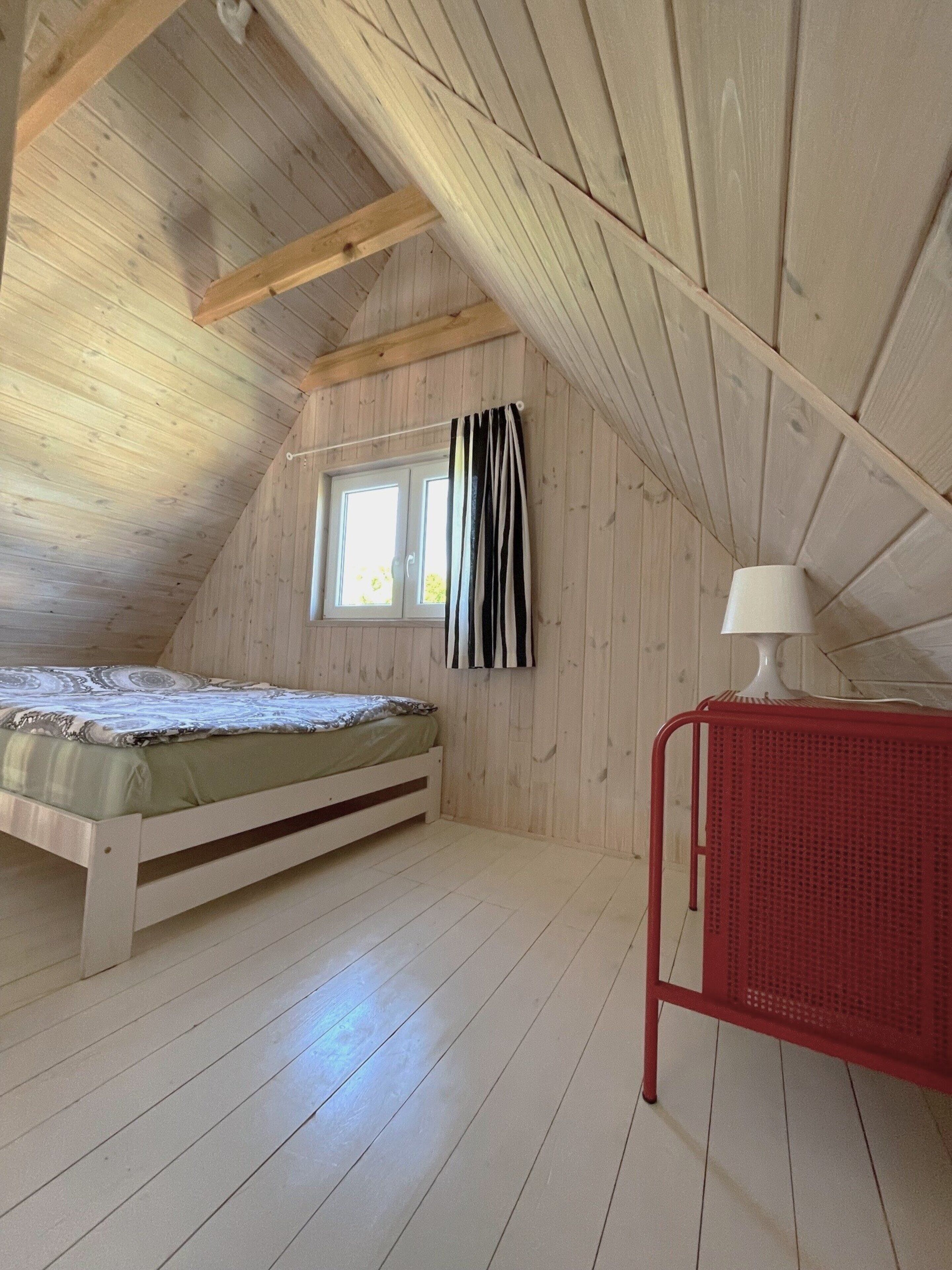 A small, sloped-roofed room with a single bed, a window with curtains, and a red metal side table with a lamp.
