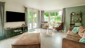 A modern living room with a flat-screen TV, a sofa, a coffee table, and a large window with a view of greenery. - Wonderful Holiday Home nearby Efteling with Private Heated Pool & Hot Tub (Kaatsheuvel)