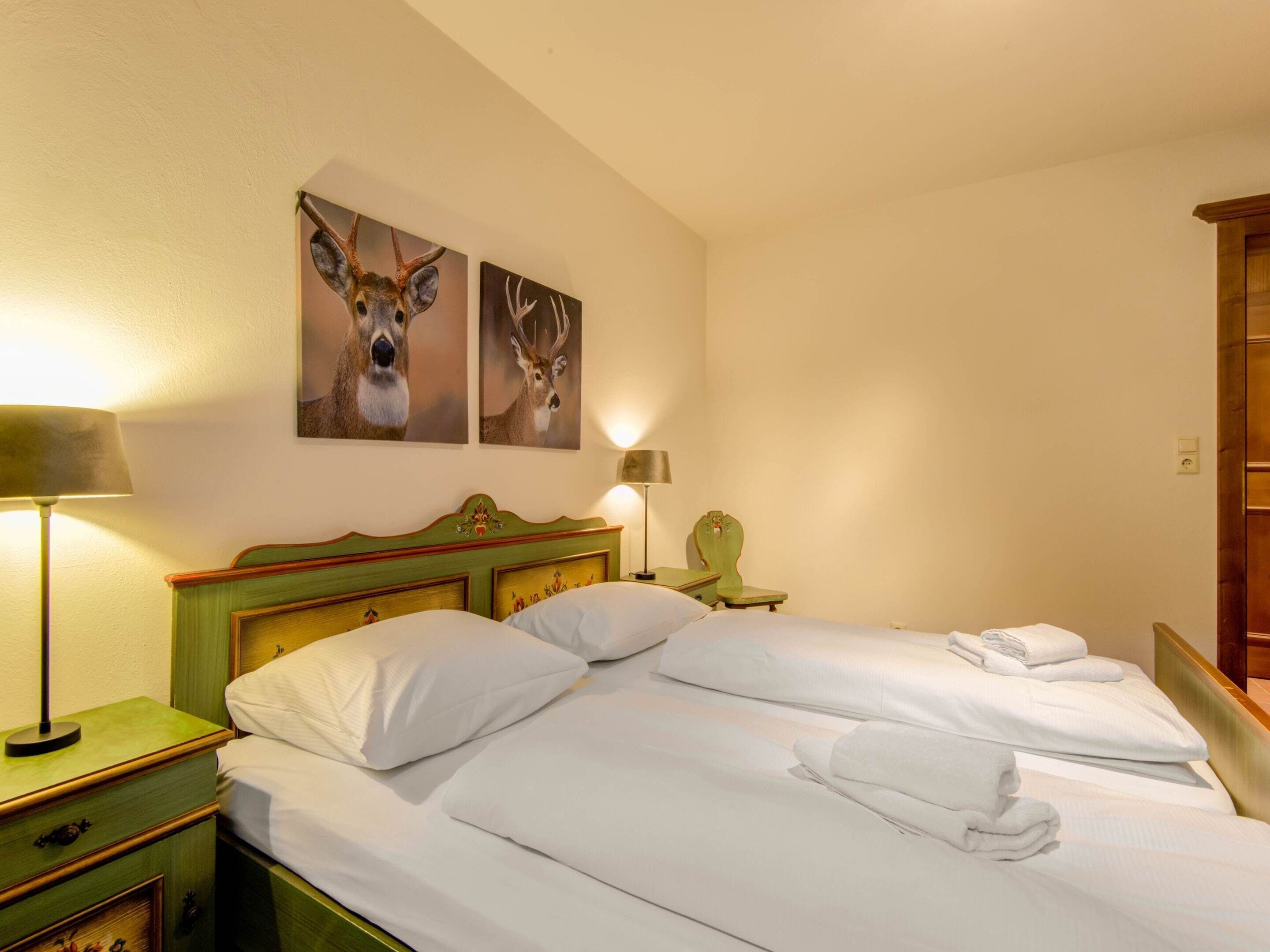 Ein Hotelzimmer mit zwei Betten, einem grünen Kopfteil, einem Nachttisch mit Lampe und zwei an der Wand hängenden Bildern von Hirschen.
