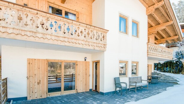 A two-story wooden chalet with a balcony, large sliding doors, and a patio area with outdoor seating.