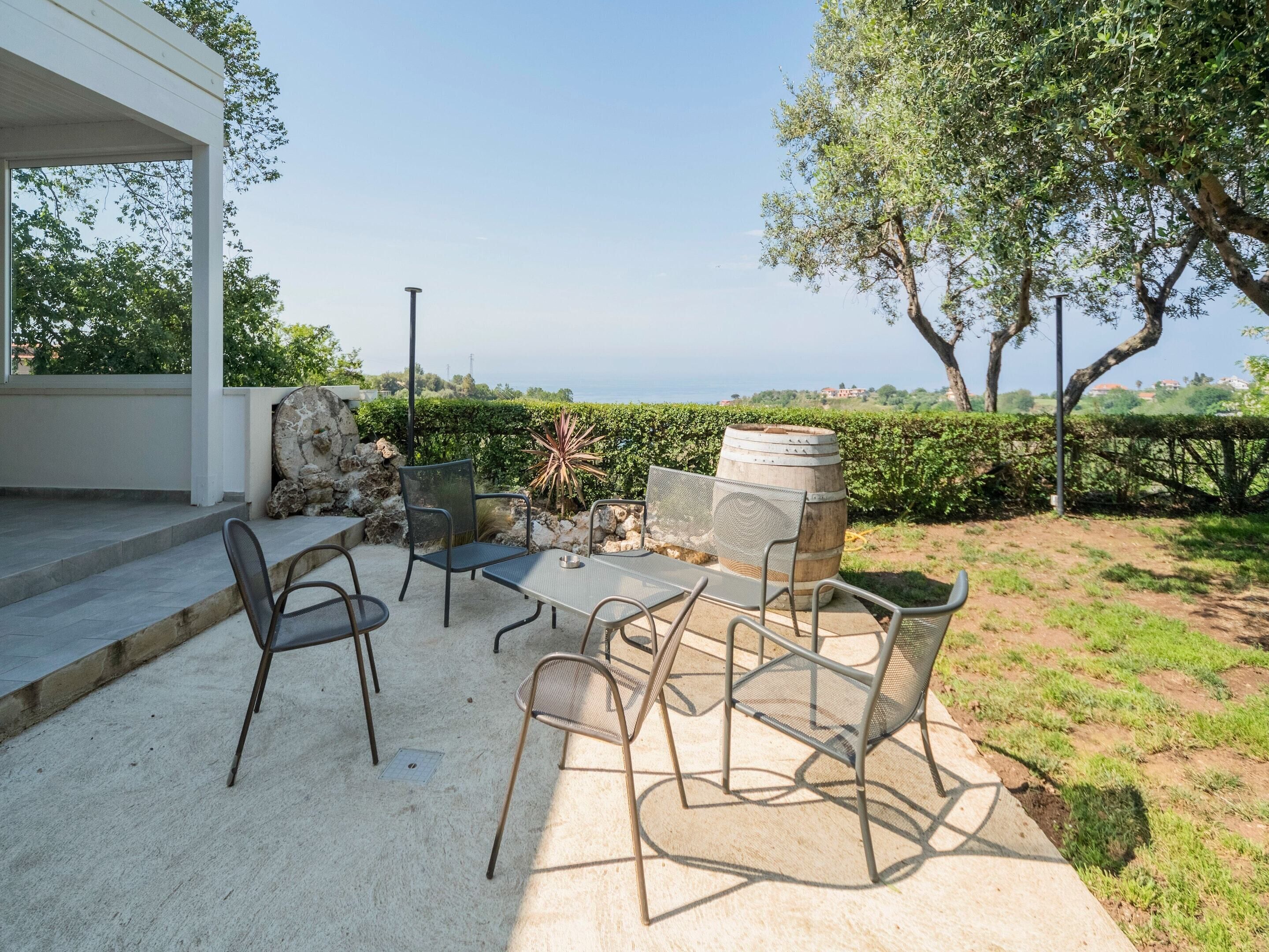 An outdoor patio with a table and chairs, a stone barrel, and a view of the sea.
