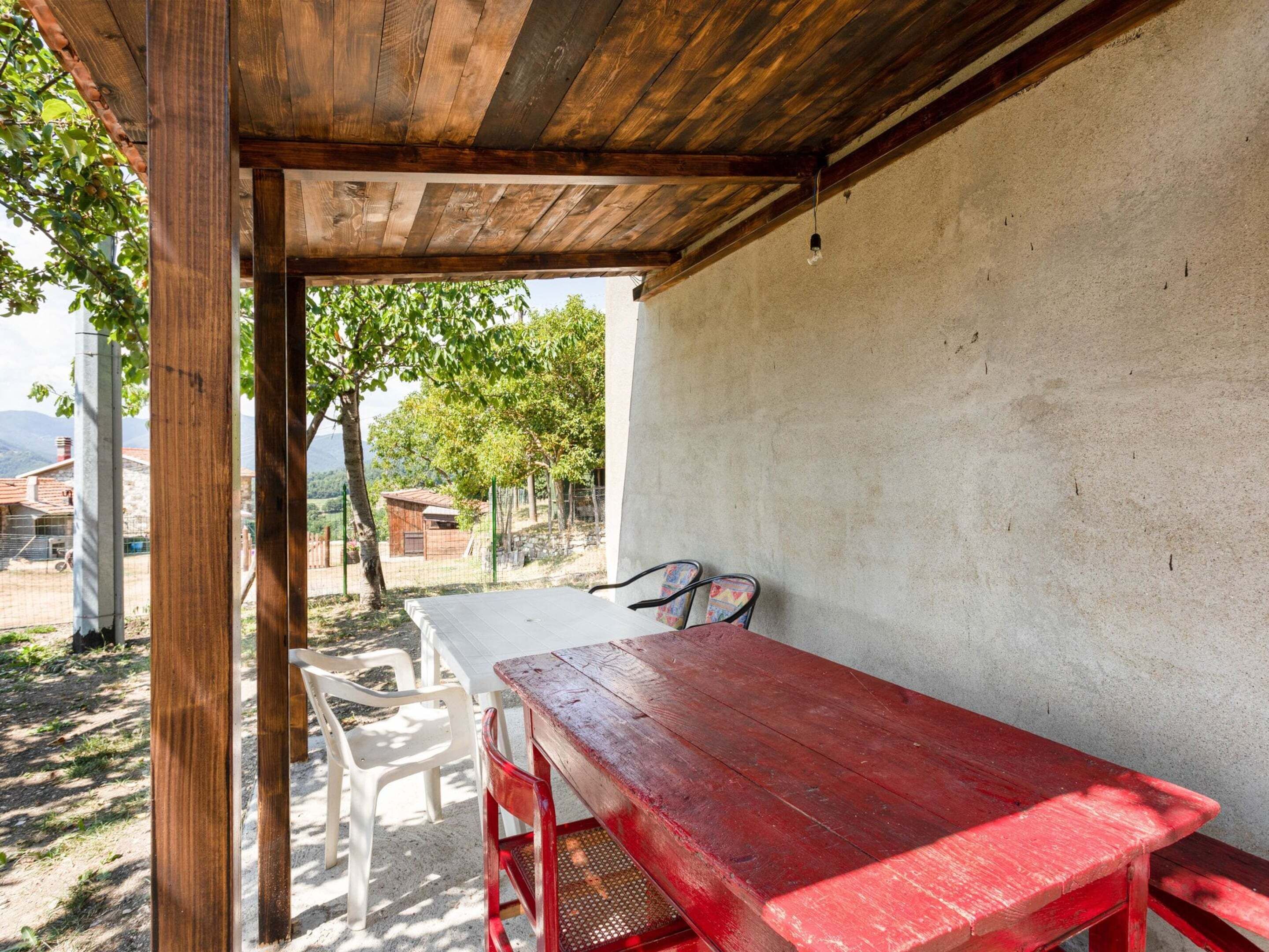 Un patio al aire libre con una mesa de madera roja y sillas blancas, un muro de concreto y vistas a árboles y edificios al fondo.