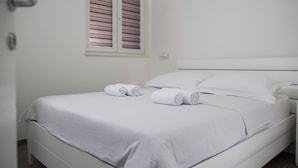 A neatly made bed with white linens and two rolled towels, a white nightstand, and a window with blinds. - Plush Apartment in Imotski near Blue & Red Lakes (Imotski)