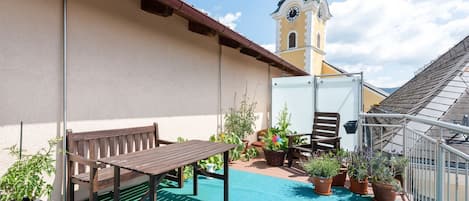A rooftop terrace with a wooden table and bench, potted plants, and a clock tower in the background.