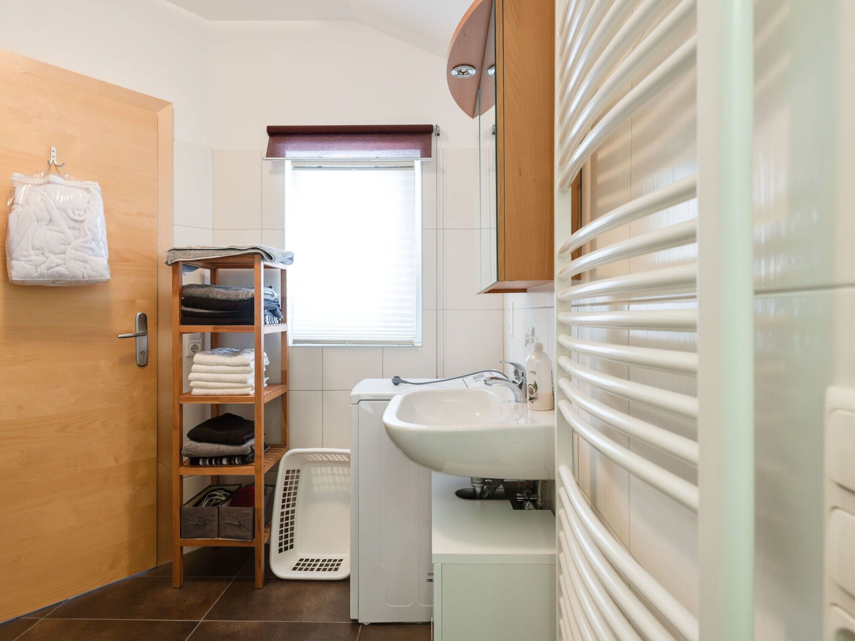 A compact bathroom with a wooden door, a shelf with folded towels, a white sink, and a window with blinds.