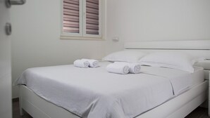 Ein ordentlich bezogenes Bett mit weißen Leinentüchern und zwei aufgerollten Handt üchern, ein weißer Nachttisch und ein Fenster mit Jalousien.