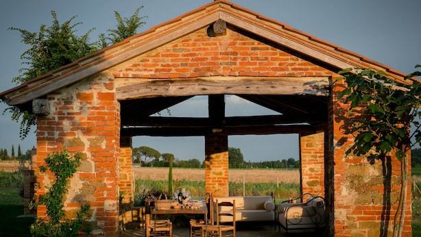 Un pavillon rustique en briques, avec une table et des chaises, entouré de verdure et avec un champ en arrière-plan.