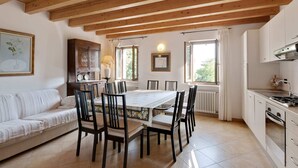 Zona pranzo con soffitto in legno, tavolo e sedie bianche. Presenza di angolo cottura con mobili bianchi e piano cottura.
