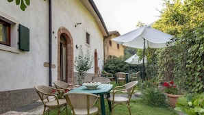 Un patio con tavolo e sedie, circondato dal verde e da un edificio con finestre ad arco.