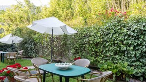 Um pátio com mesa e cadeiras, cercado por vegetação exuberante e um guarda-sol branco.
