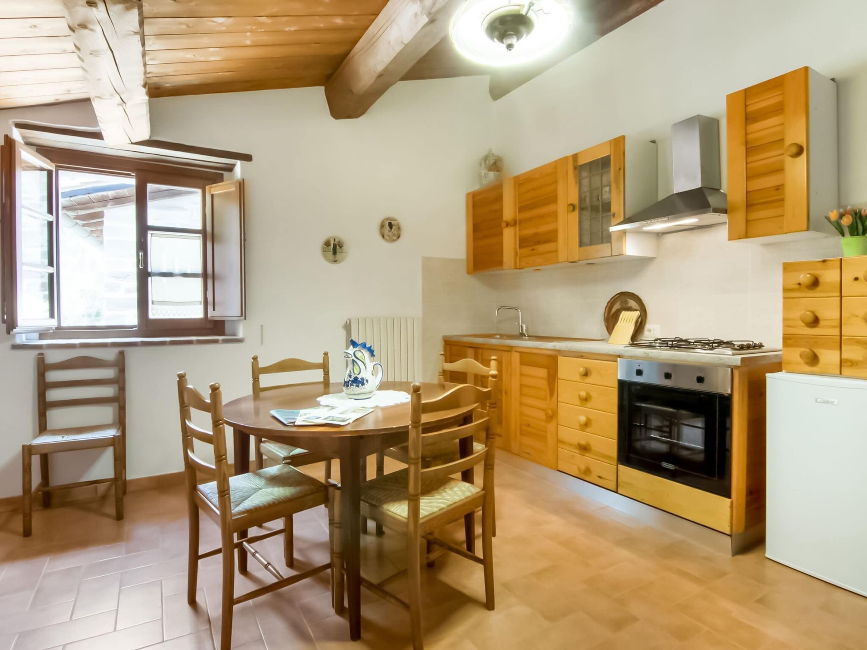 Une cuisine avec des meubles de rangement en bois, une cuisinière et un réfrigérateur. On y trouve une table à manger avec des chaises et une fenêtre avec des stores.