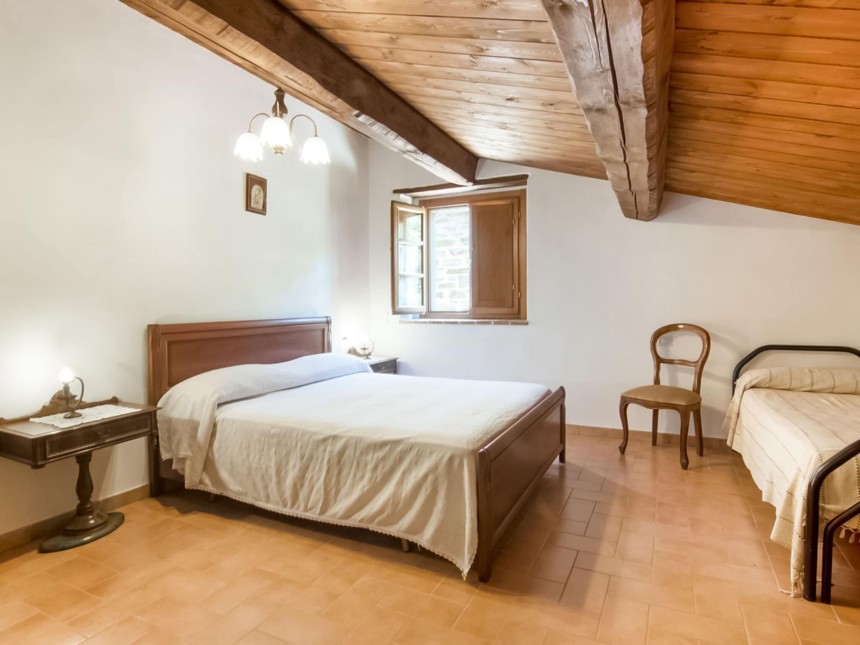A bedroom with a wooden ceiling, a bed with a wooden frame, a bedside table with a lamp, a chair, and a window with a view.