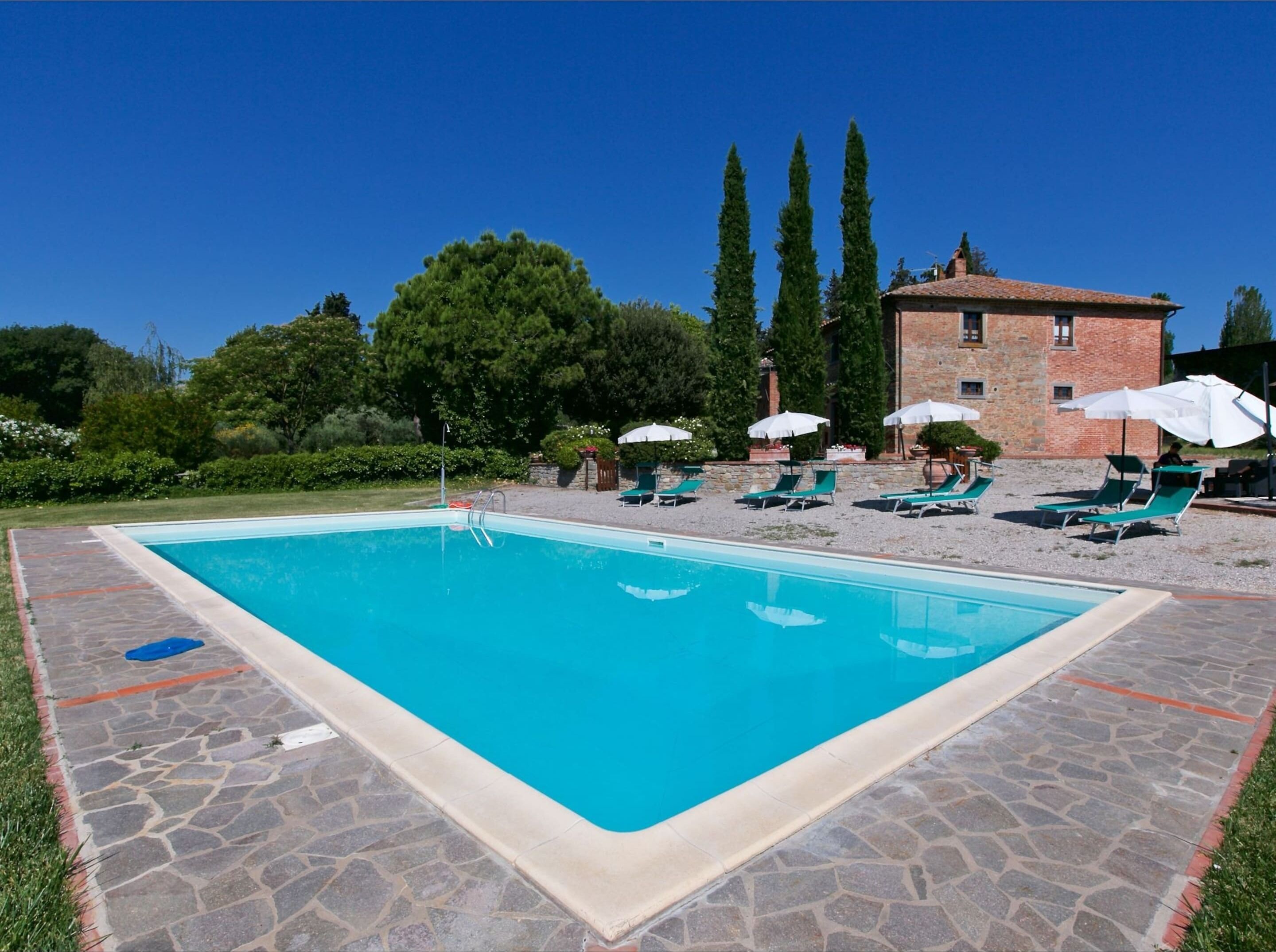 Una piscina rectangular con borde de piedra, rodeada de sillas de descanso y sombrillas, con un edificio y árboles al fondo.