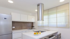 Een moderne keuken met witte kasten, een roestvrijstalen koelkast, een centraal kookeiland met barkrukken en een raam met jaloezieën.