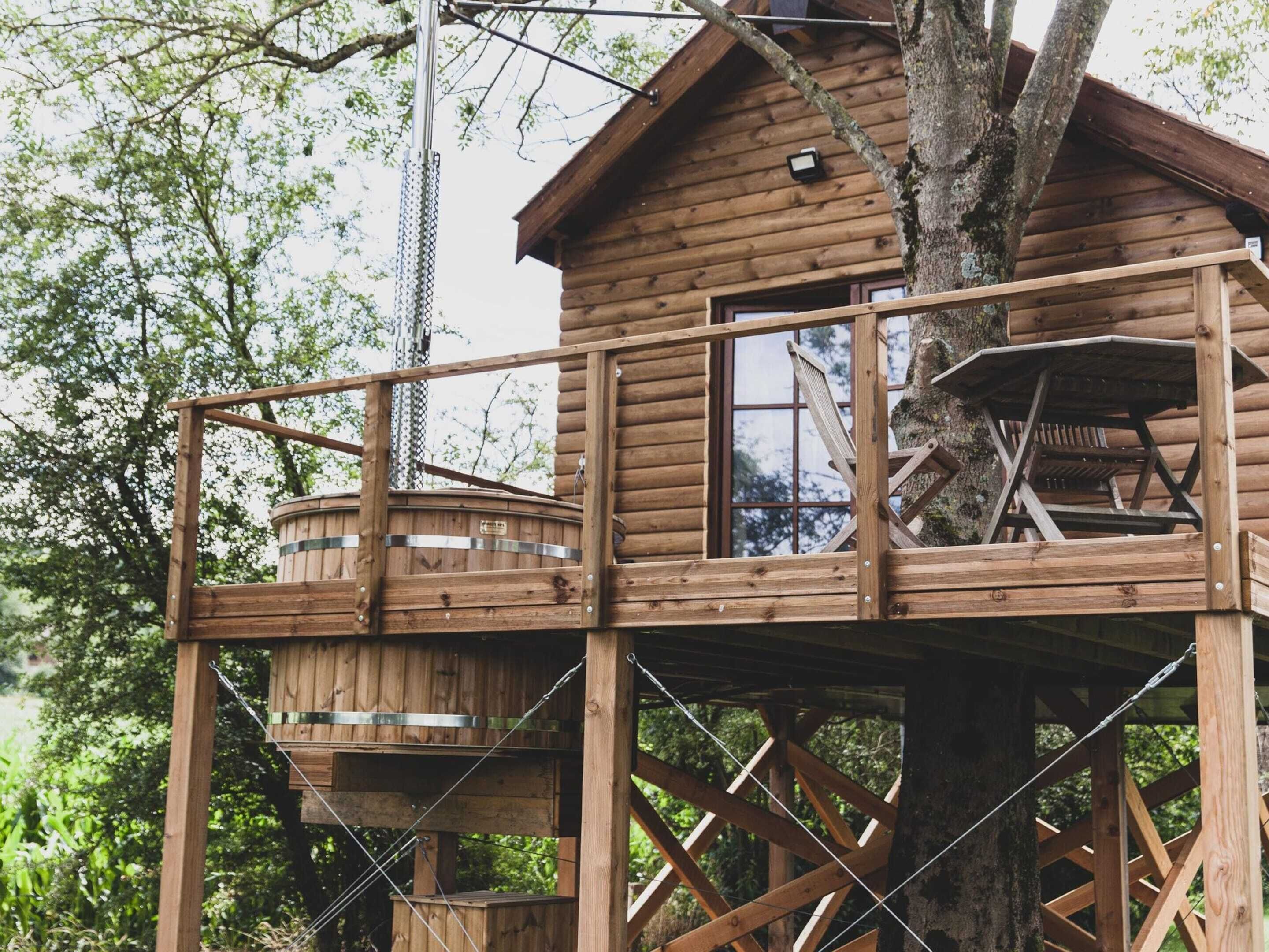 A wooden treehouse with a balcony and a lower level, surrounded by trees.