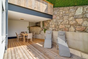 A wooden deck with a table and chairs, a lounge chair, and a stone wall.
