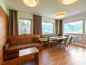 A modern room with a wooden floor, a brown leather sofa, a wooden table with chairs, and large windows offering a mountain view.