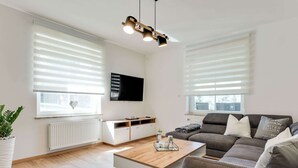 Una sala de estar moderna con un sofá, una mesa de centro y un televisor montado en la pared. Hay ventanas amplias con persianas, un radiador y una planta en maceta.