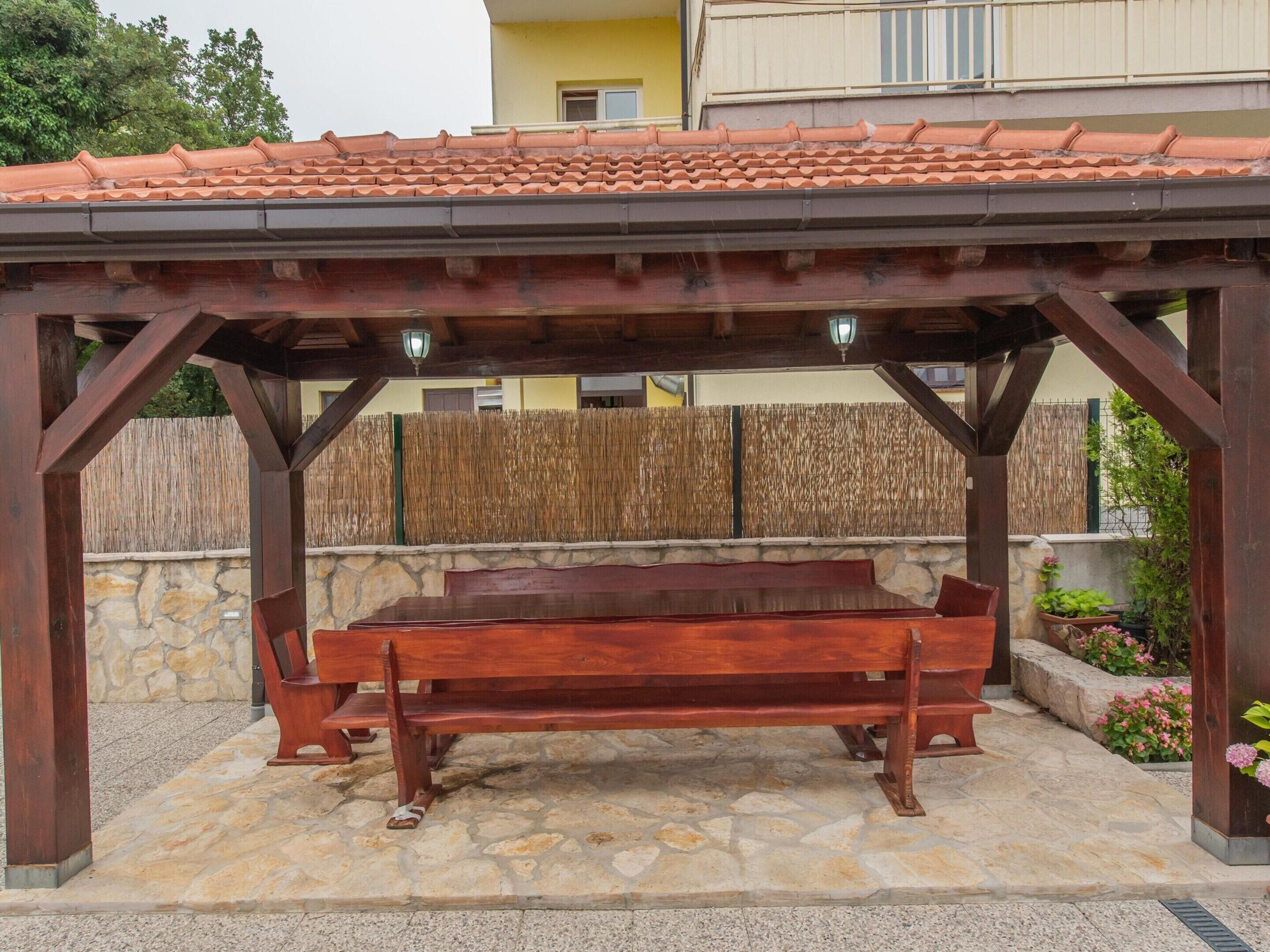 A wooden pergola with a bench underneath, stone flooring, and a backdrop of a building and a fence.