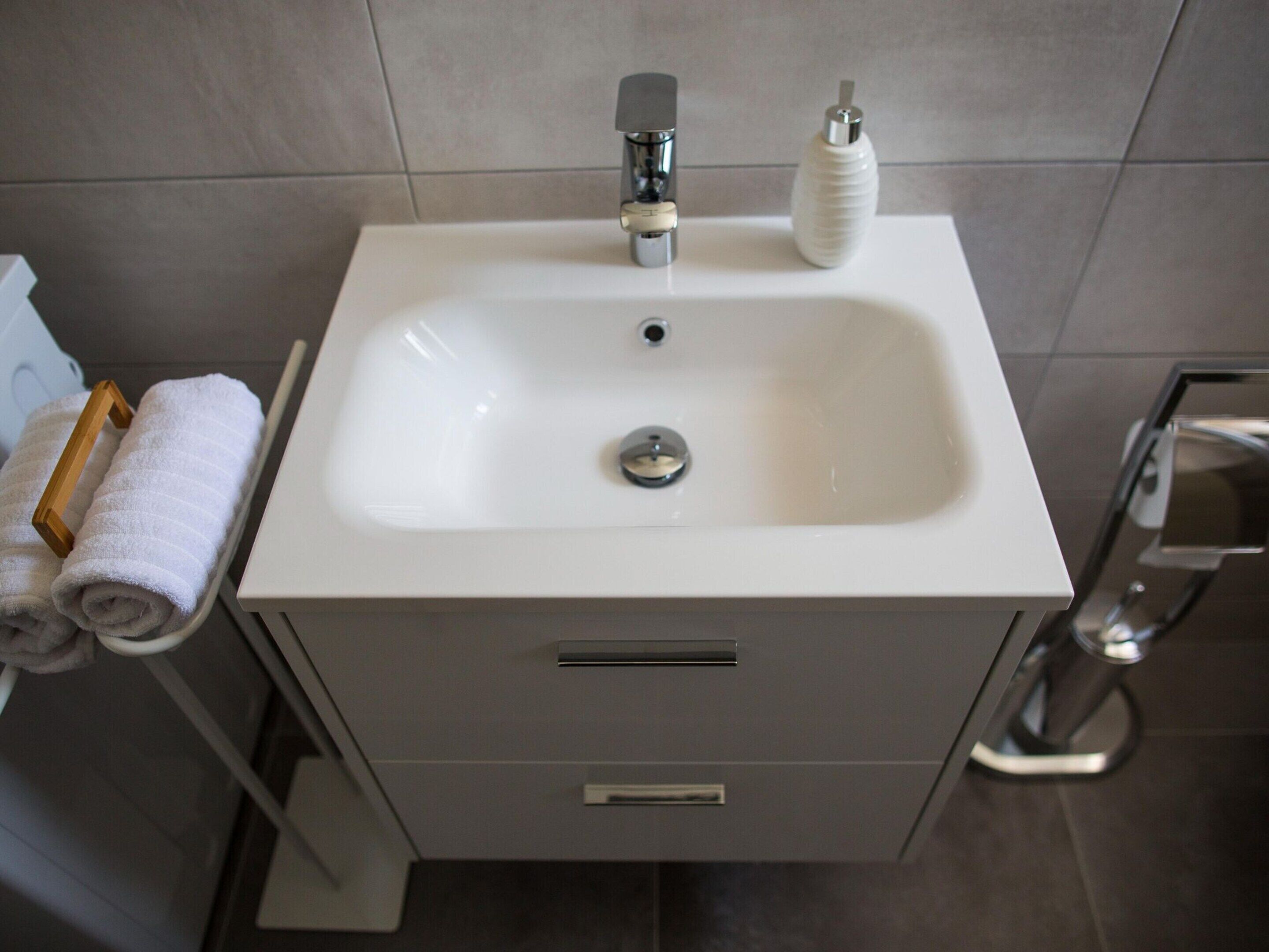 A modern bathroom with a white sink, chrome faucet, and a soap dispenser. There are two towels on a rack and a shower chair to the right.