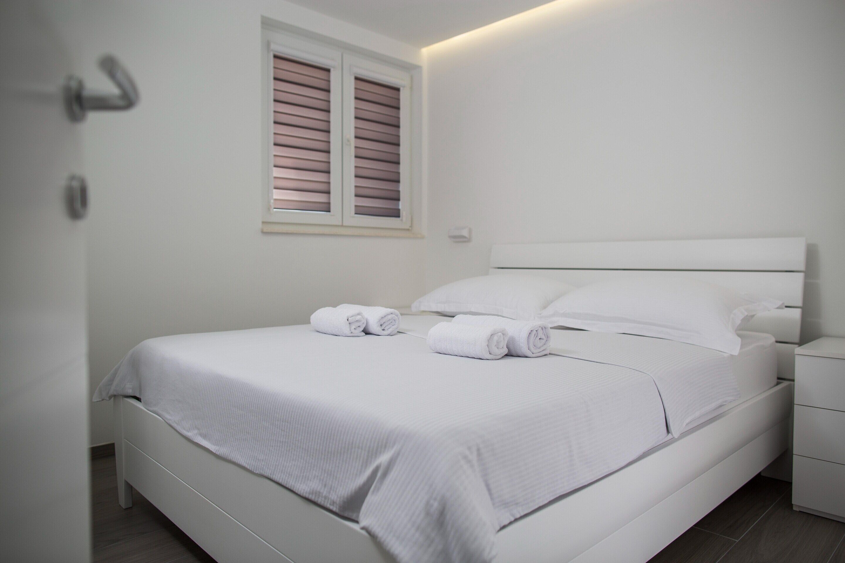A neatly made bed with white linens and pillows, a bedside table, and a window with blinds.