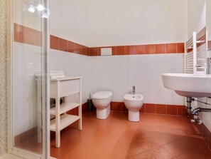 A bathroom with white and orange tiles, a glass shower enclosure, a toilet, a bidet, and a sink.