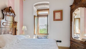 A bedroom with a large bed, a wooden dresser, a mirror, a window with a view, and a small table with a lamp.