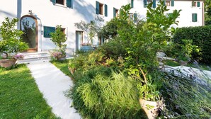 A white building with green shutters, a stone staircase, and a well-maintained garden with various plants and a pathway.