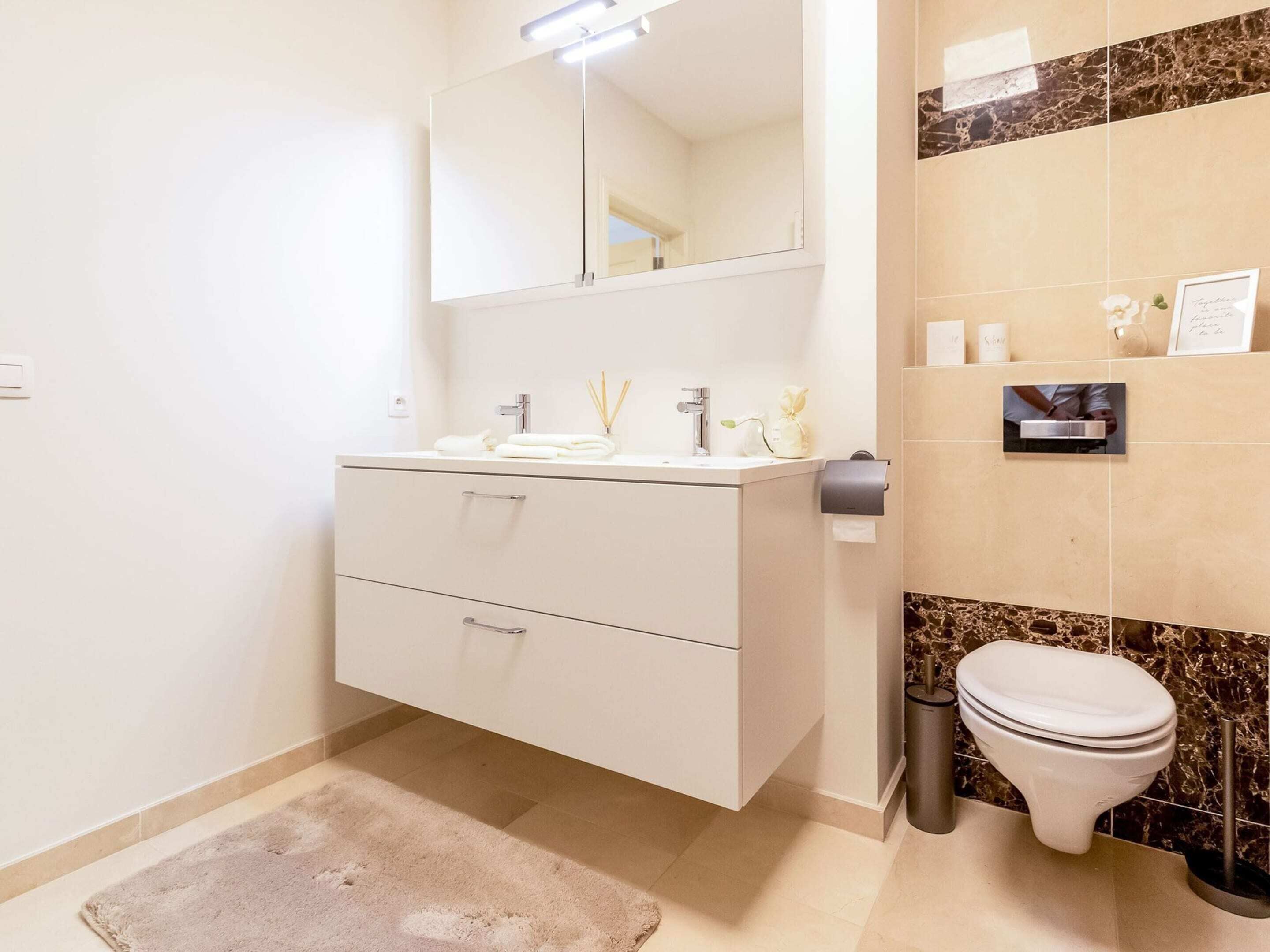 A modern bathroom with a vanity, mirror, toilet, and marble backsplash.