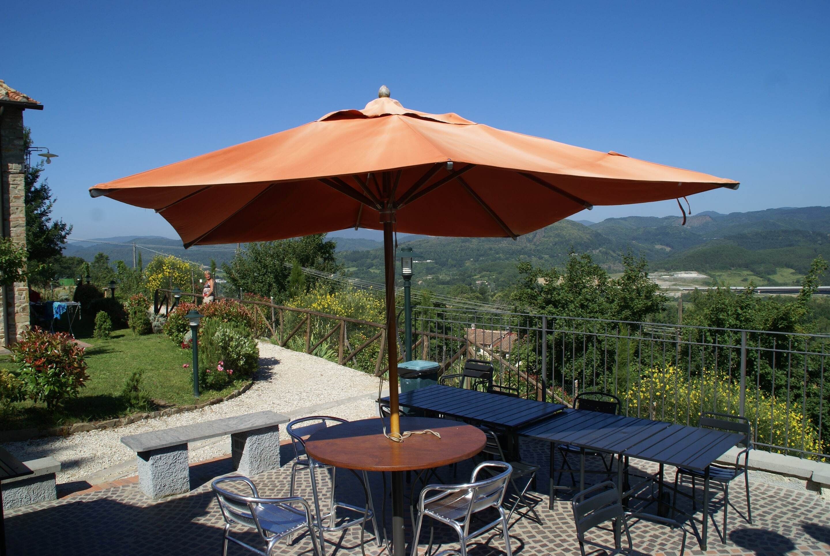 A patio with a table, chairs, and an umbrella, overlooking a garden and distant hills.